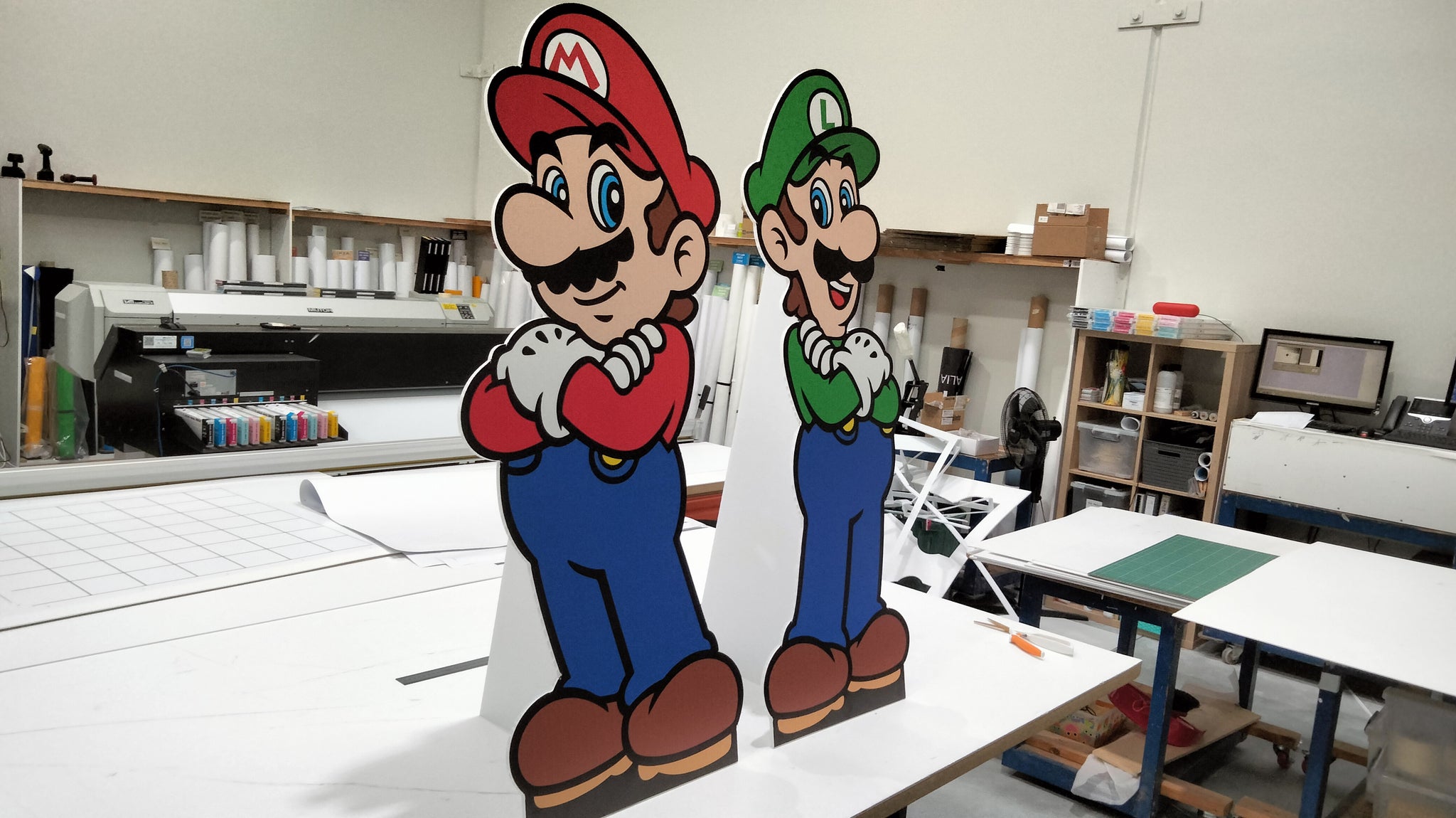 Two People Cutouts | Standees | Cut to Shape Displays of cartoon characters are showcased on a print shop table amid printing equipment—perfect inspiration for custom event signage and corflute signs.