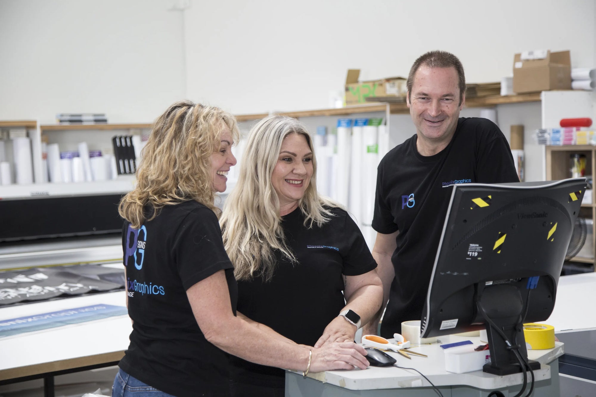 Three people in black shirts with a logo, working at a desk with a computer in an office setting.